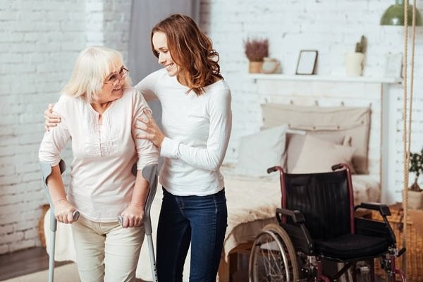 I will help you. Positive aged disabled woman waling with crutches while her caring smiling granddaughter helping her at home