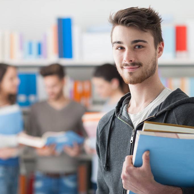 Confident student posing in the library Confident handsome student holding books and smiling at camera, library bookshelves on background, learning and education concept